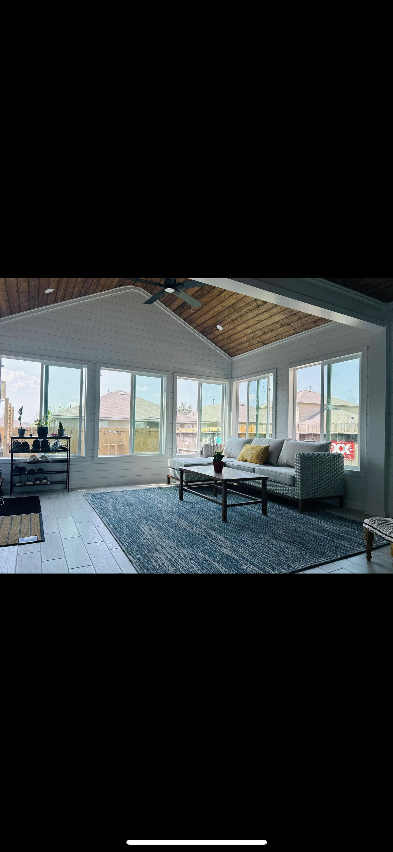 Sunlit enclosed porch with white walls, large windows, a wooden ceiling with a ceiling fan, a white couch with yellow and white pillows, a coffee table, a blue area rug, a black shoe rack with shoes and plants, and a view of neighboring houses.