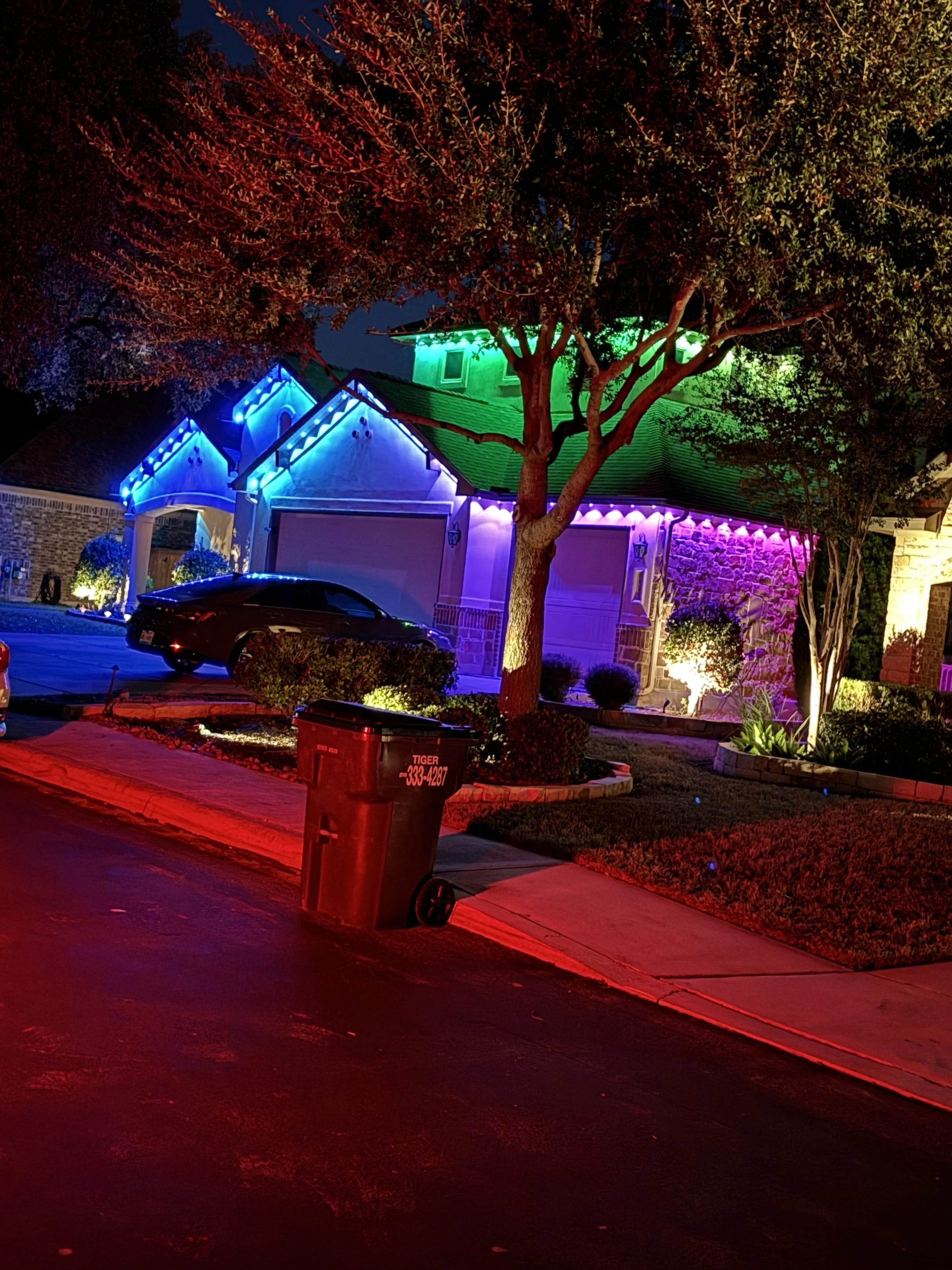 House decorated with colorful rainbow LED lights at night, with a large tree in the front yard, a black car parked in driveway, and a red trash bin on the sidewalk.