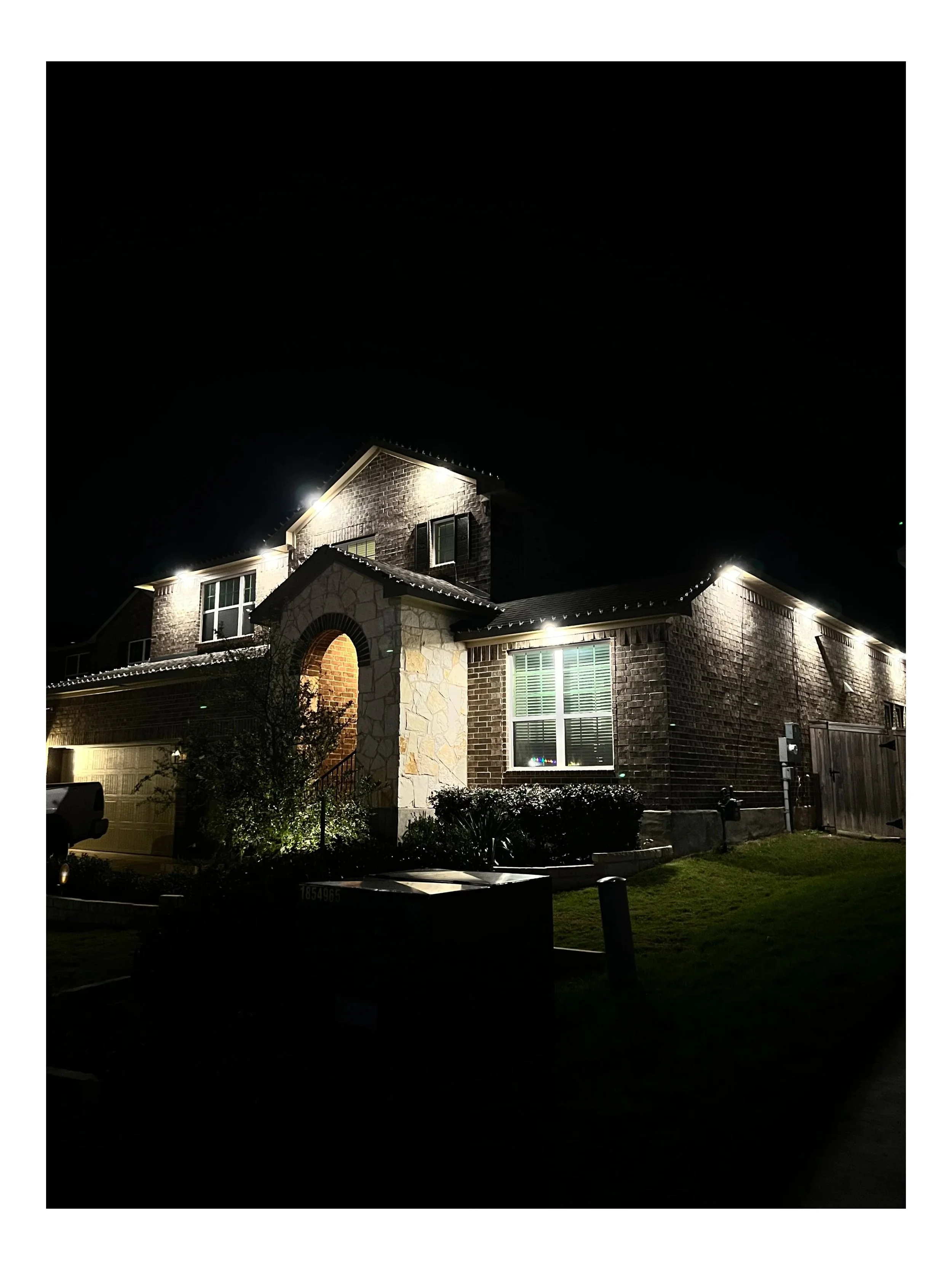 A two-story brick house at night with exterior lights illuminating the front entrance and windows, a front lawn with bushes, and a parked vehicle partially visible on the left.