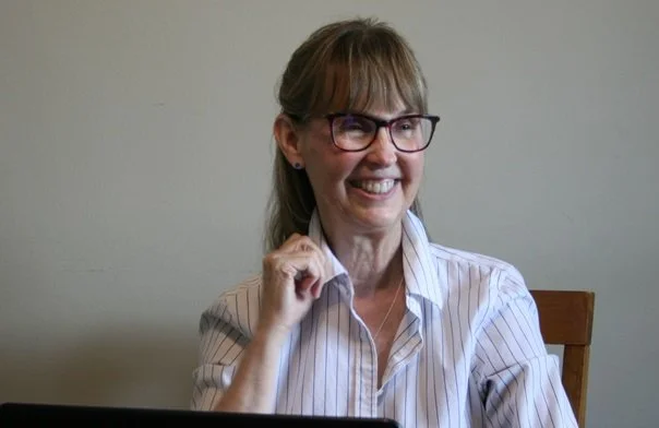 A woman with brown hair, glasses, and a striped shirt smiling and sitting at a desk.