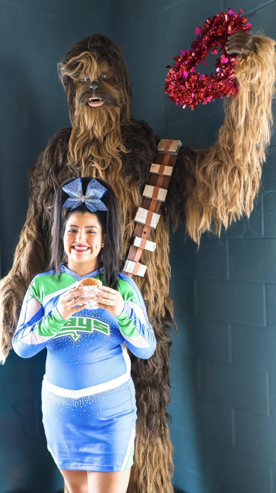 A girl in a cheerleading uniform holding a snack and smiling, standing in front of a person in a Chewbacca costume from Star Wars, who is holding a heart-shaped decoration made of red tinsel.