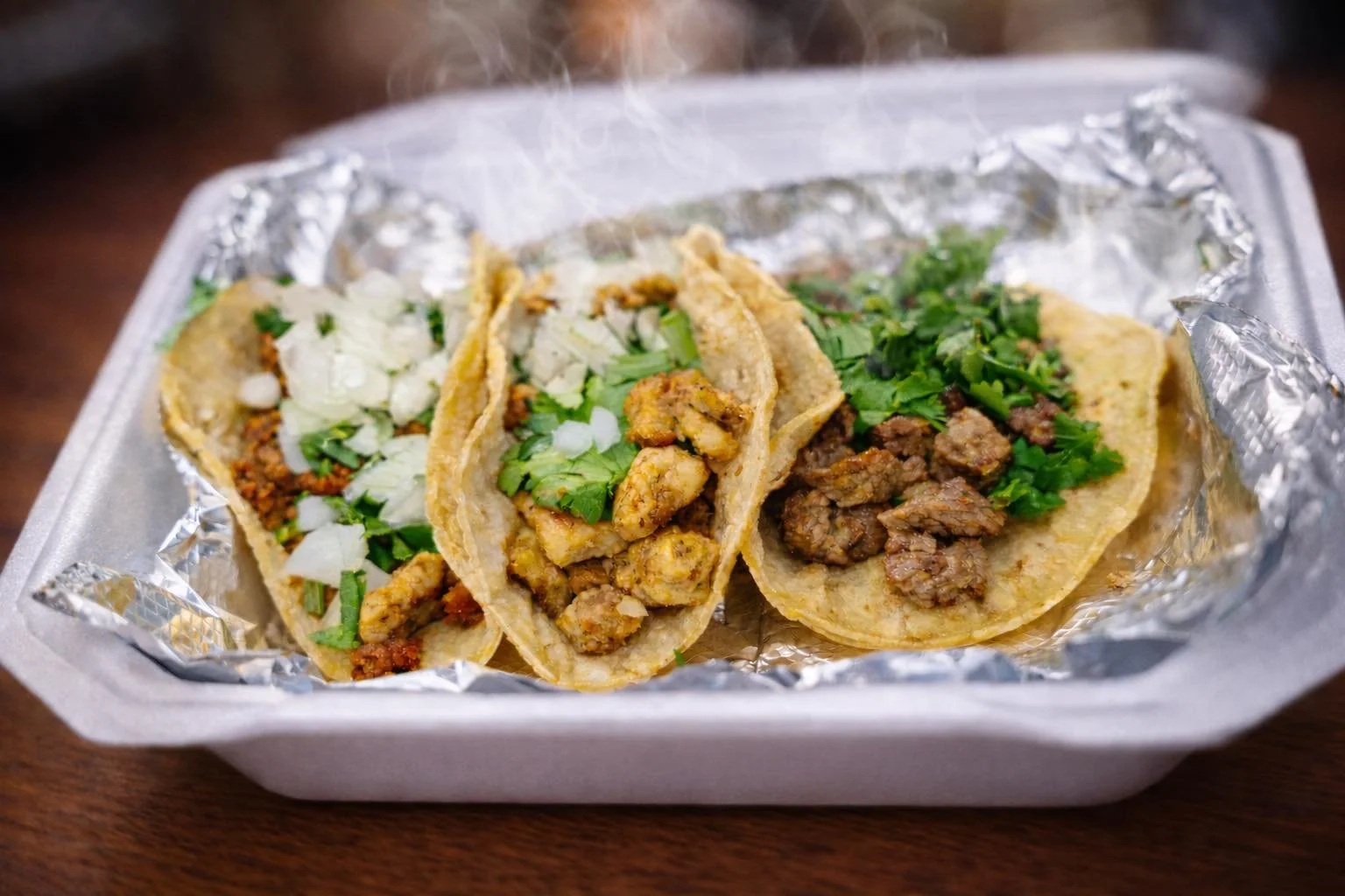Three tacos in a styrofoam container with foil, filled with meat, chopped onions, cilantro, and possibly some cheese, on a wooden surface.