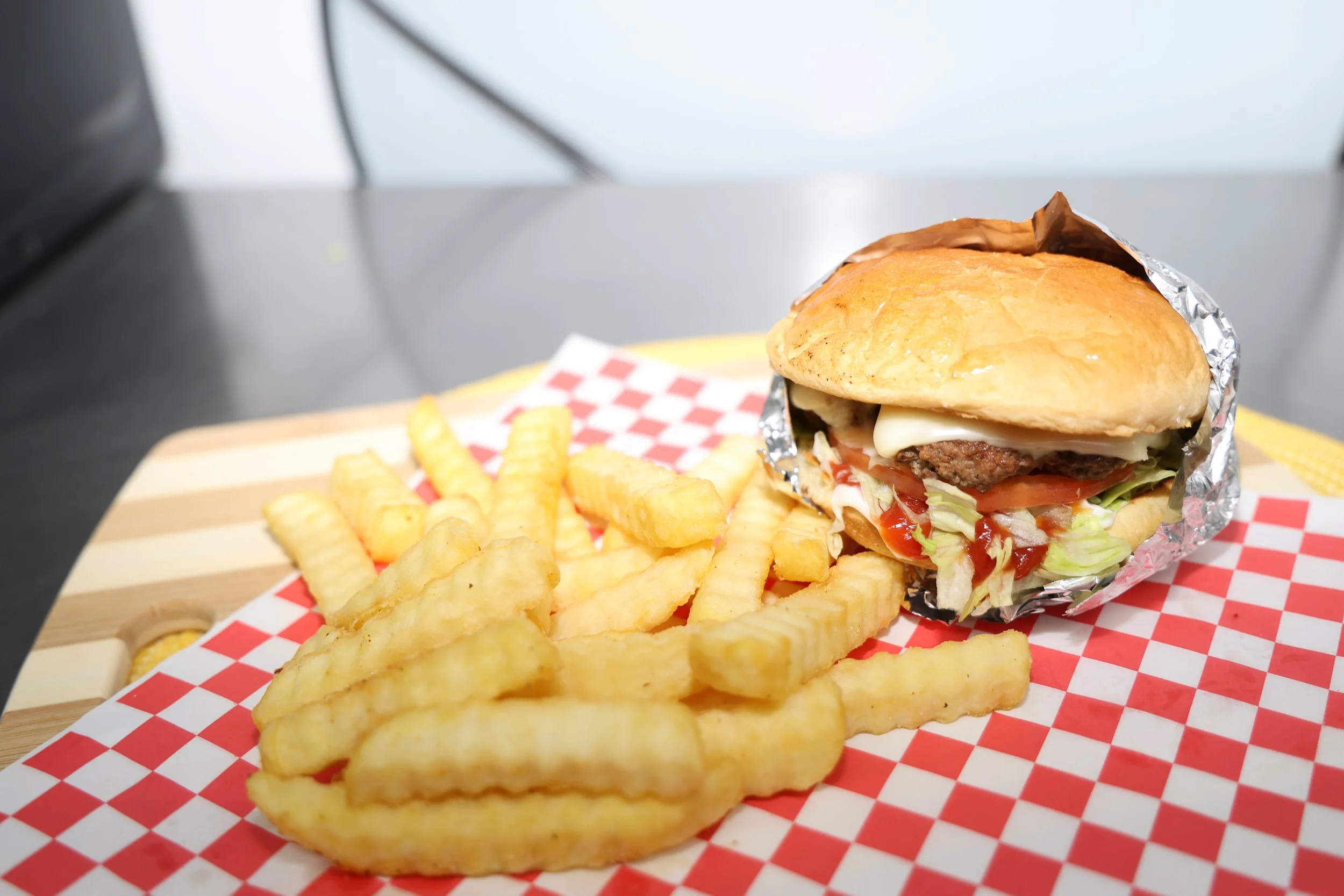A cheeseburger with lettuce, tomato, cheese, beef patty, in a bun wrapped with foil, served with crinkle-cut French fries on red and white checkered paper.