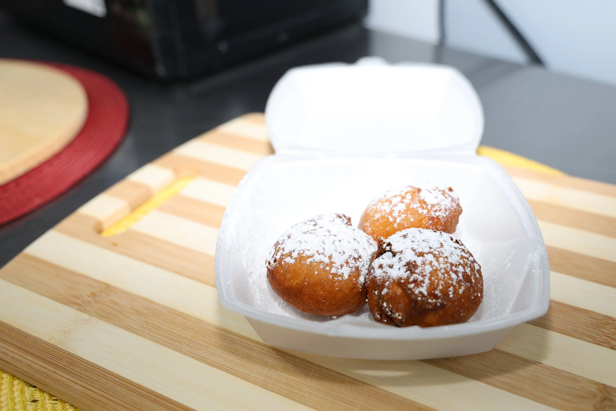 Three glazed donuts with powdered sugar in a foam takeout container on a wooden cutting board.