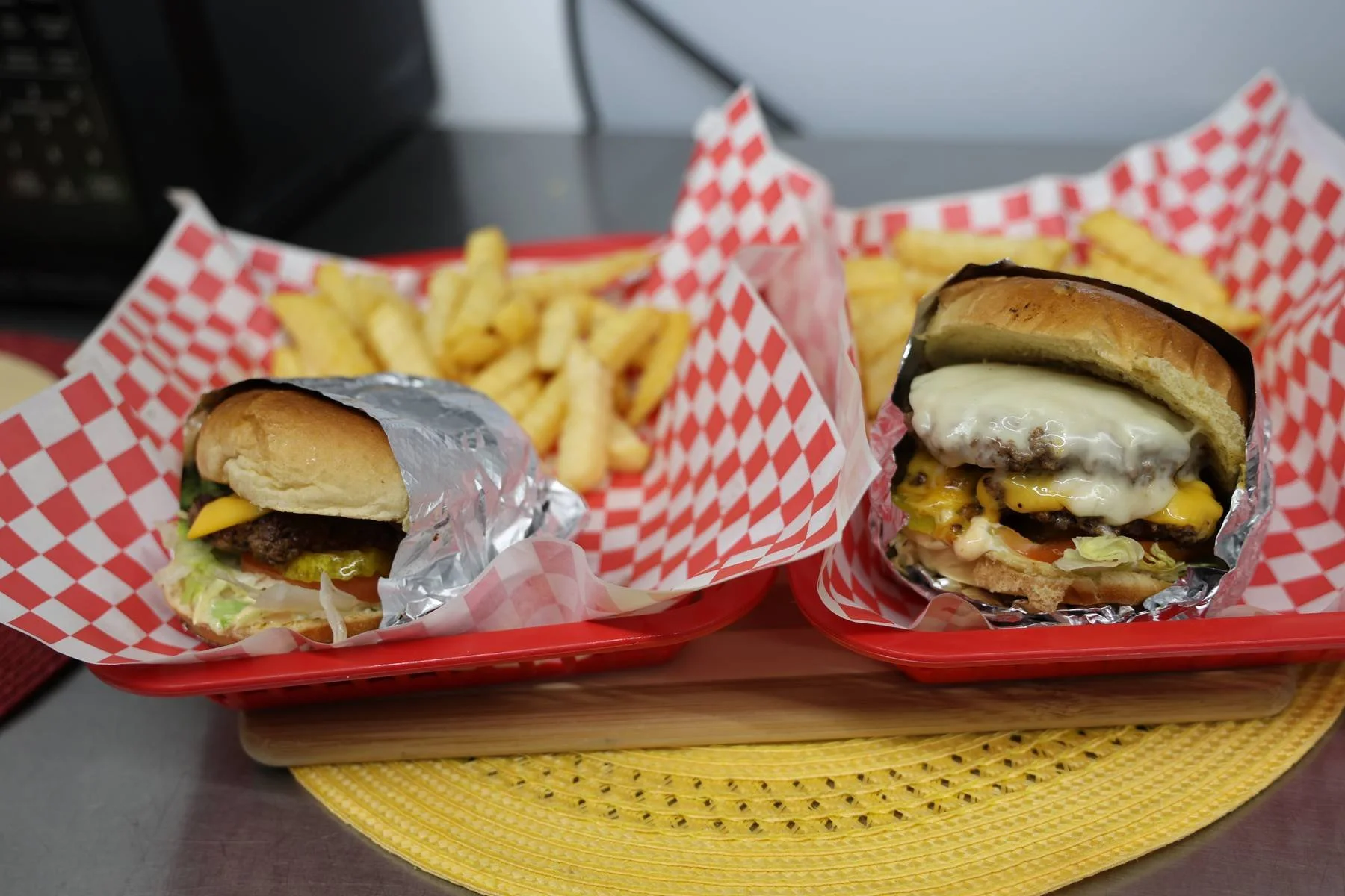 Two cheeseburgers with fries served on red trays lined with red and white checkered paper, placed on a yellow woven placemat.