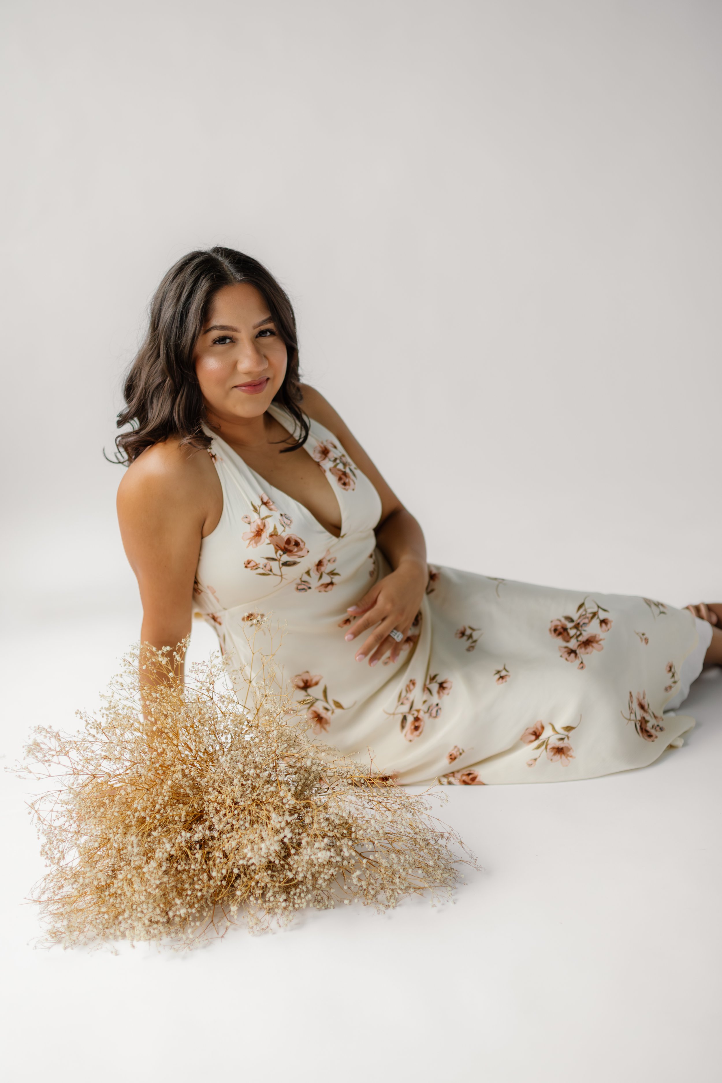 A woman with wavy dark hair in a floral cream dress, sitting on the floor with a bouquet of dried flowers, against a white background.