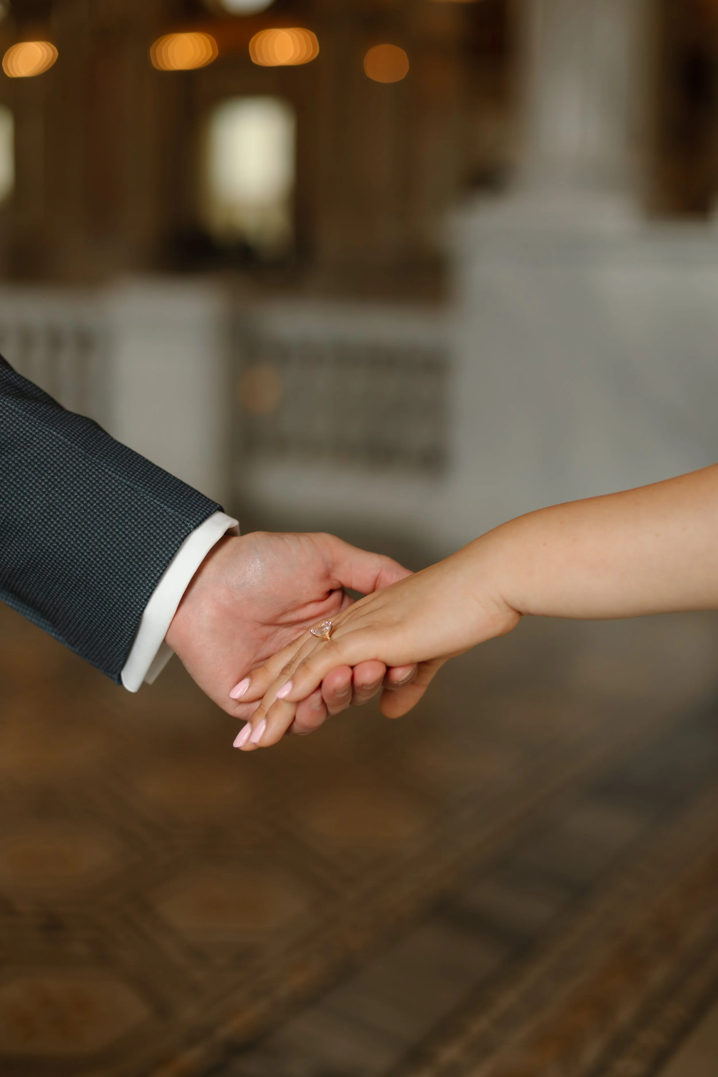 Close-up of a man in a dark suit and a woman with a ring on her finger holding hands, standing on a patterned wooden floor, with blurred lights and furniture in the background.