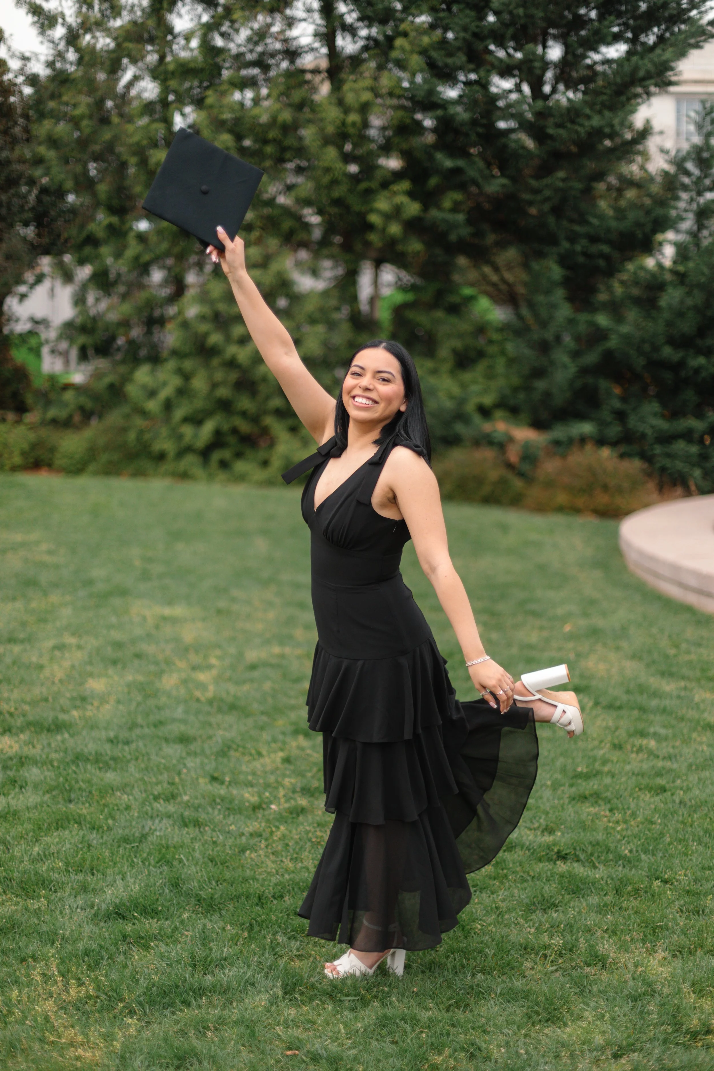 A young woman in a black dress is celebrating her graduation. She is smiling, holding her graduation cap in one hand raised in the air, and lifting her leg to show her white high heels.