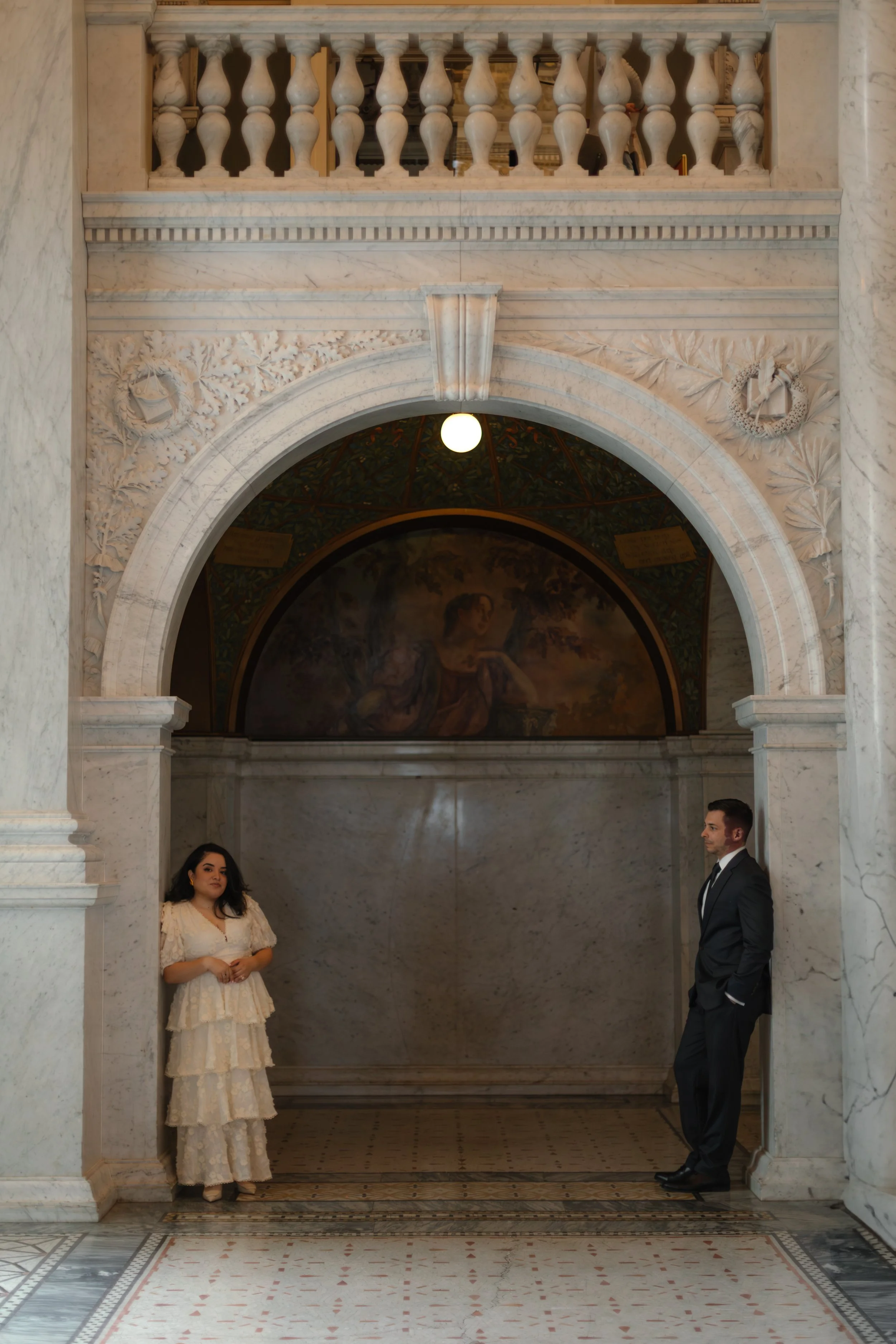 A woman in a cream-colored, layered dress and a man in a dark suit stand apart in a marble hallway with ornate architectural details, a painting behind them, and a small balcony above.