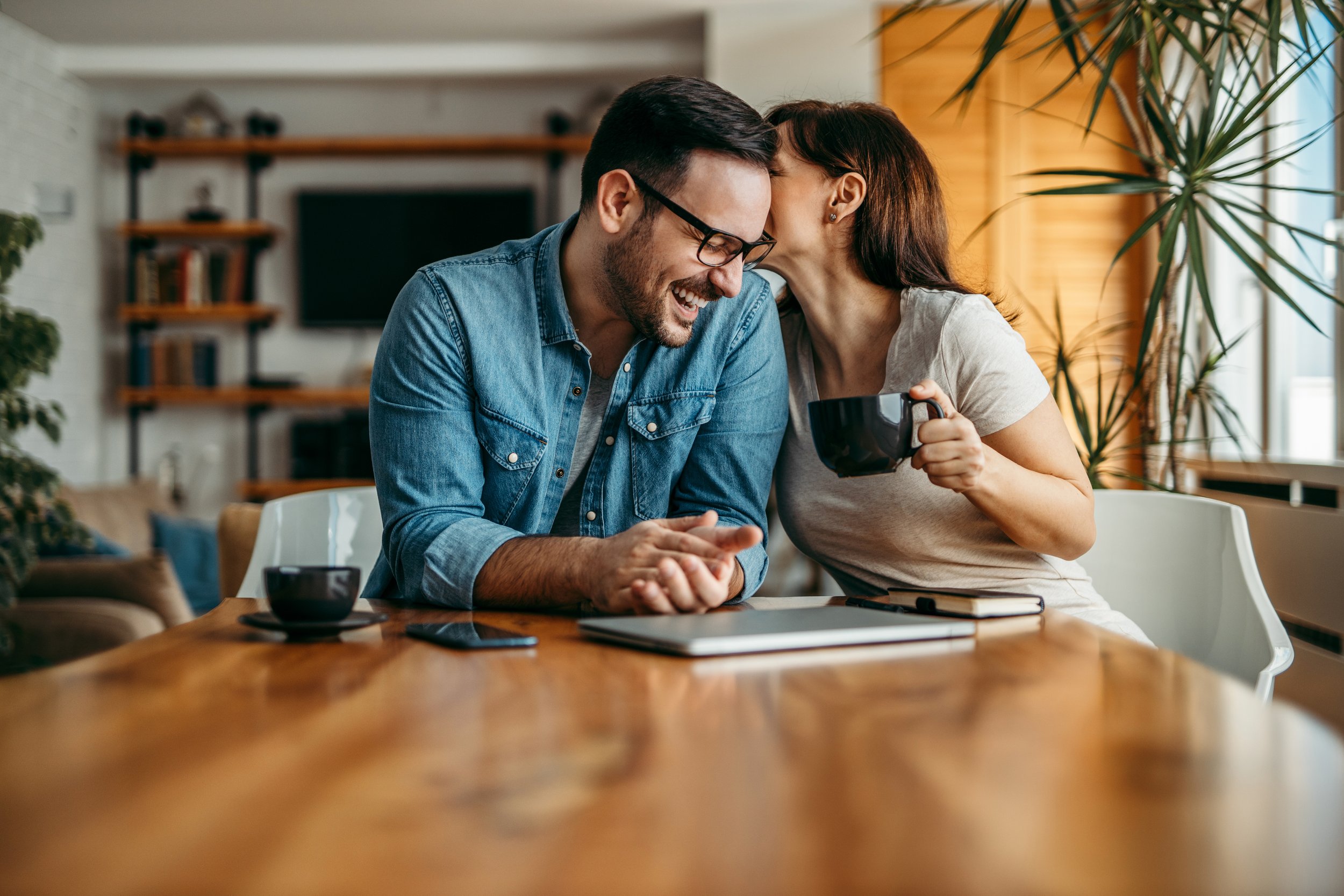 A man and woman sharing a laugh at a wooden table, with the woman whispering into the man's ear and holding a black mug, in a cozy living room.