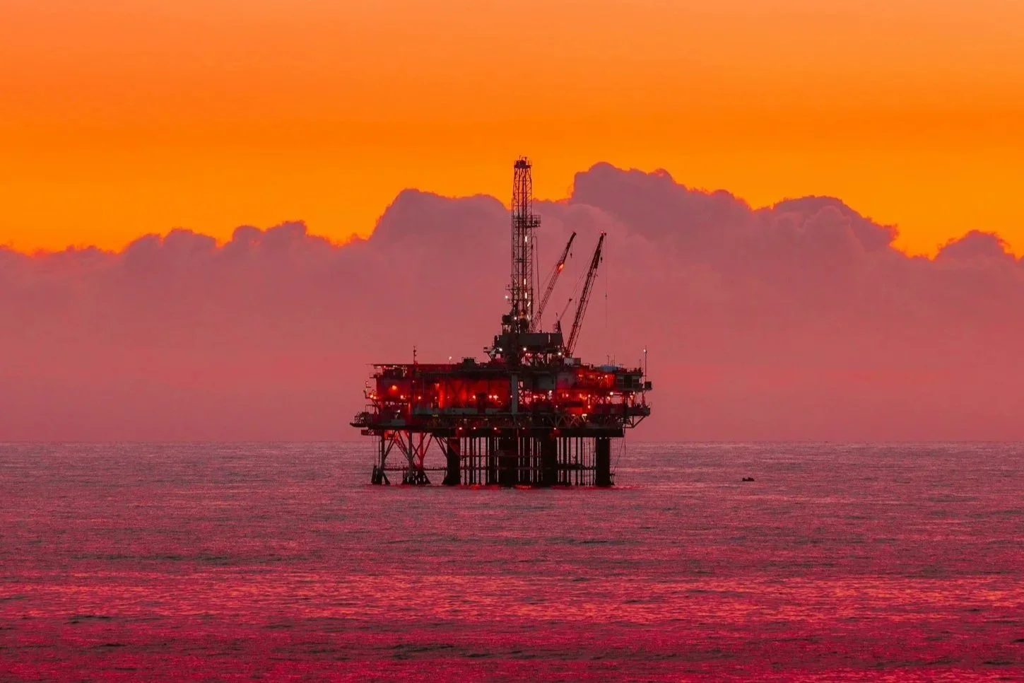 Offshore oil rig in the ocean during sunset with a vibrant orange and pink sky and illuminated lights on the rig.