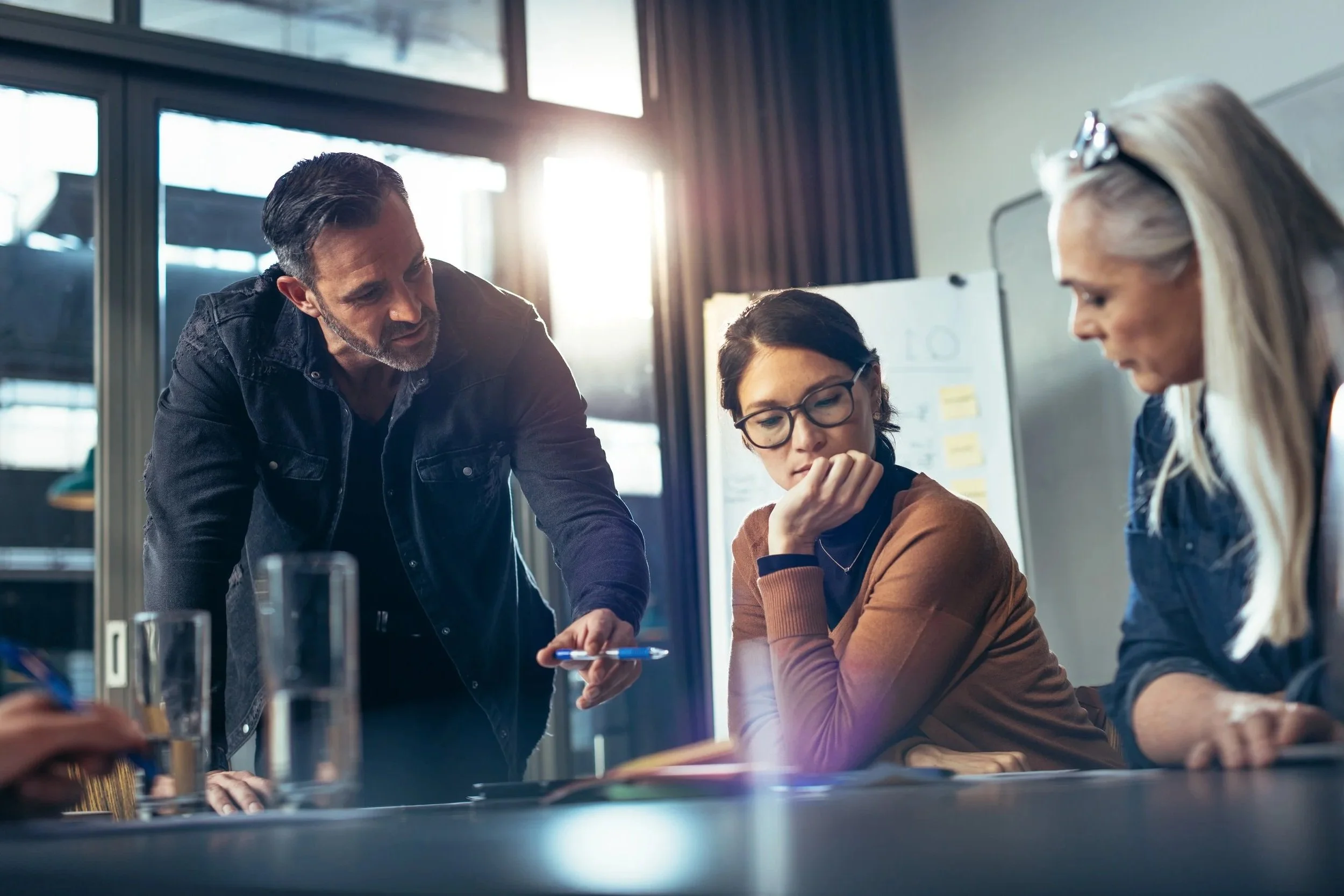 Four people having a serious discussion around a table in a well-lit office, with a man standing and pointing at a document, two women seated listening and contemplating.