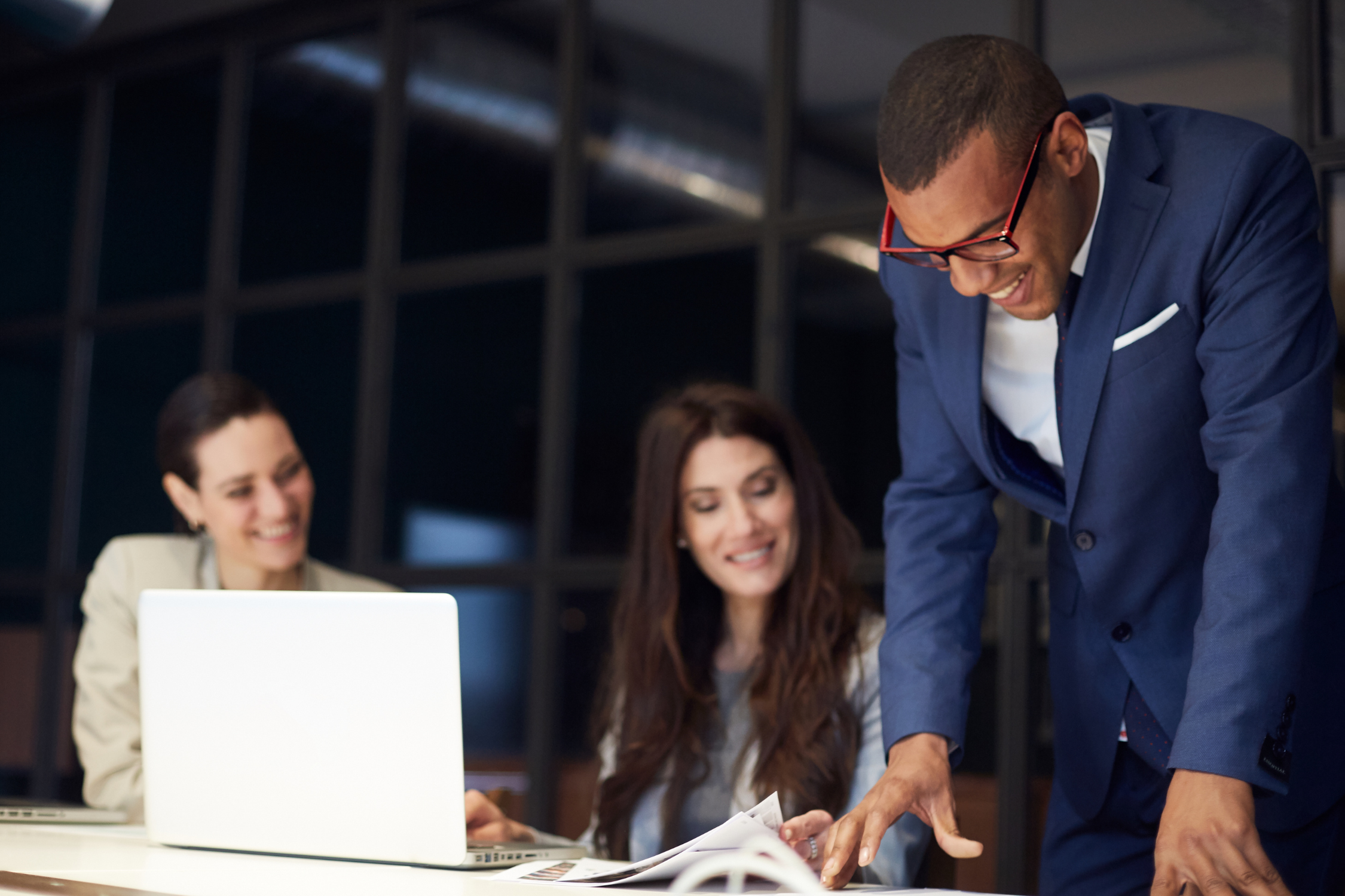 Three diverse business professionals in a meeting, two women and one man, are smiling and listening as a man in a blue suit shows or reviews documents at a conference table with a laptop.