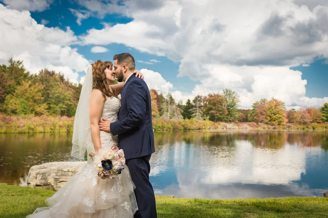 Bride and groom embracing beside a lake with trees and cloudy sky in the background.