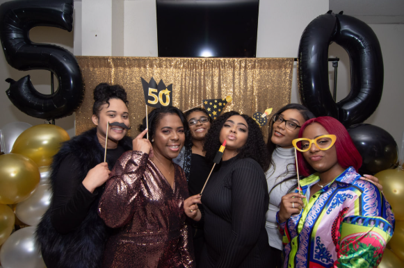 Group of six women celebrating a 50th birthday with gold and black balloons, some wearing festive accessories and holding photo props.