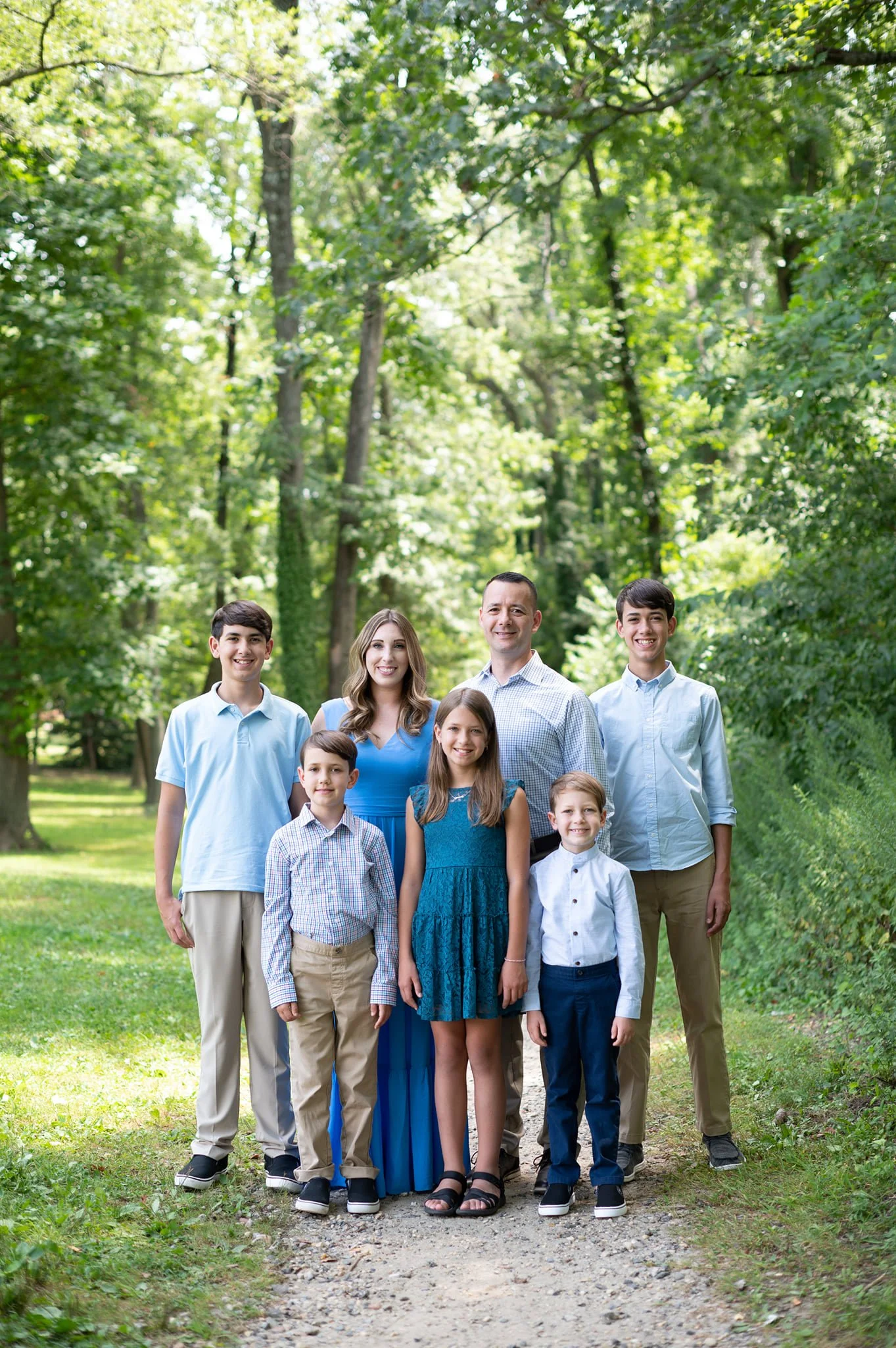 A family of seven standing outdoors on a dirt path in a lush green park, smiling at the camera.