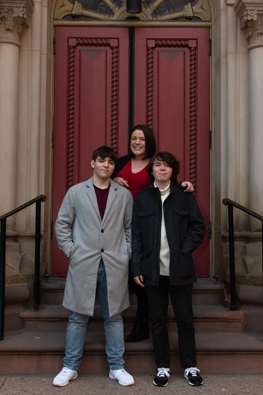 A woman stands between two teenage boys on the steps of a building with large pink double doors. The woman is smiling, wearing a red top and black pants. One boy wears a gray coat and jeans, the other a black jacket and black pants. They are outdoors in front of the building.