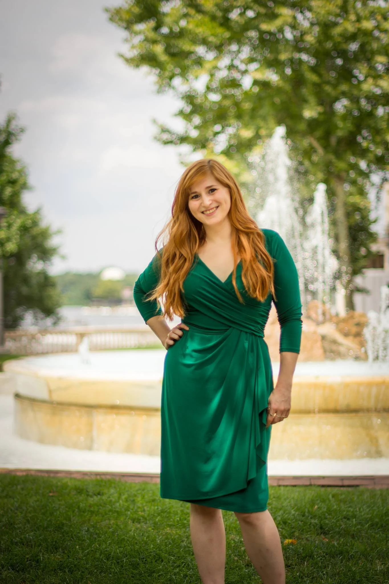 A woman with long red hair wearing a green dress, standing outdoors in front of a fountain and trees, smiling at the camera.