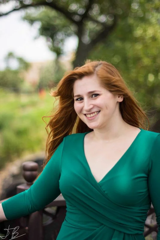 A woman with long red hair smiling outdoors in a green dress with a V-neckline in a park setting with trees and grass.