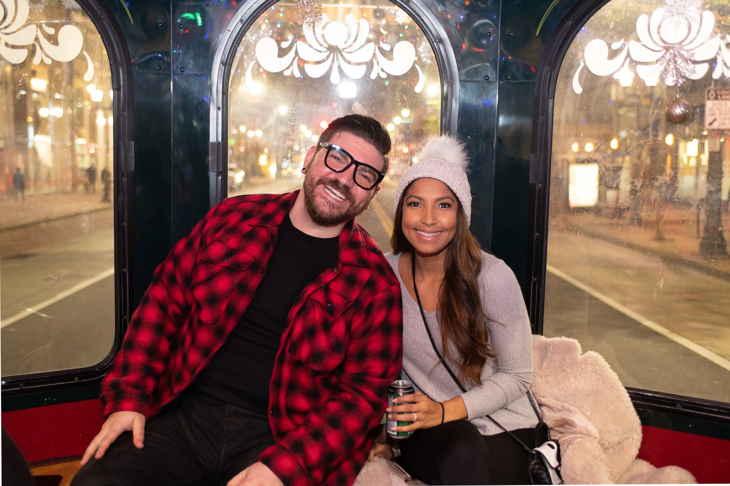 A smiling man and woman sit inside a decorated vehicle during the evening, with blurred city lights outside. The man wears glasses, a black shirt, and a red and black plaid jacket. The woman wears a gray sweater, a knit beanie with a pom-pom, and holds a can of beverage.