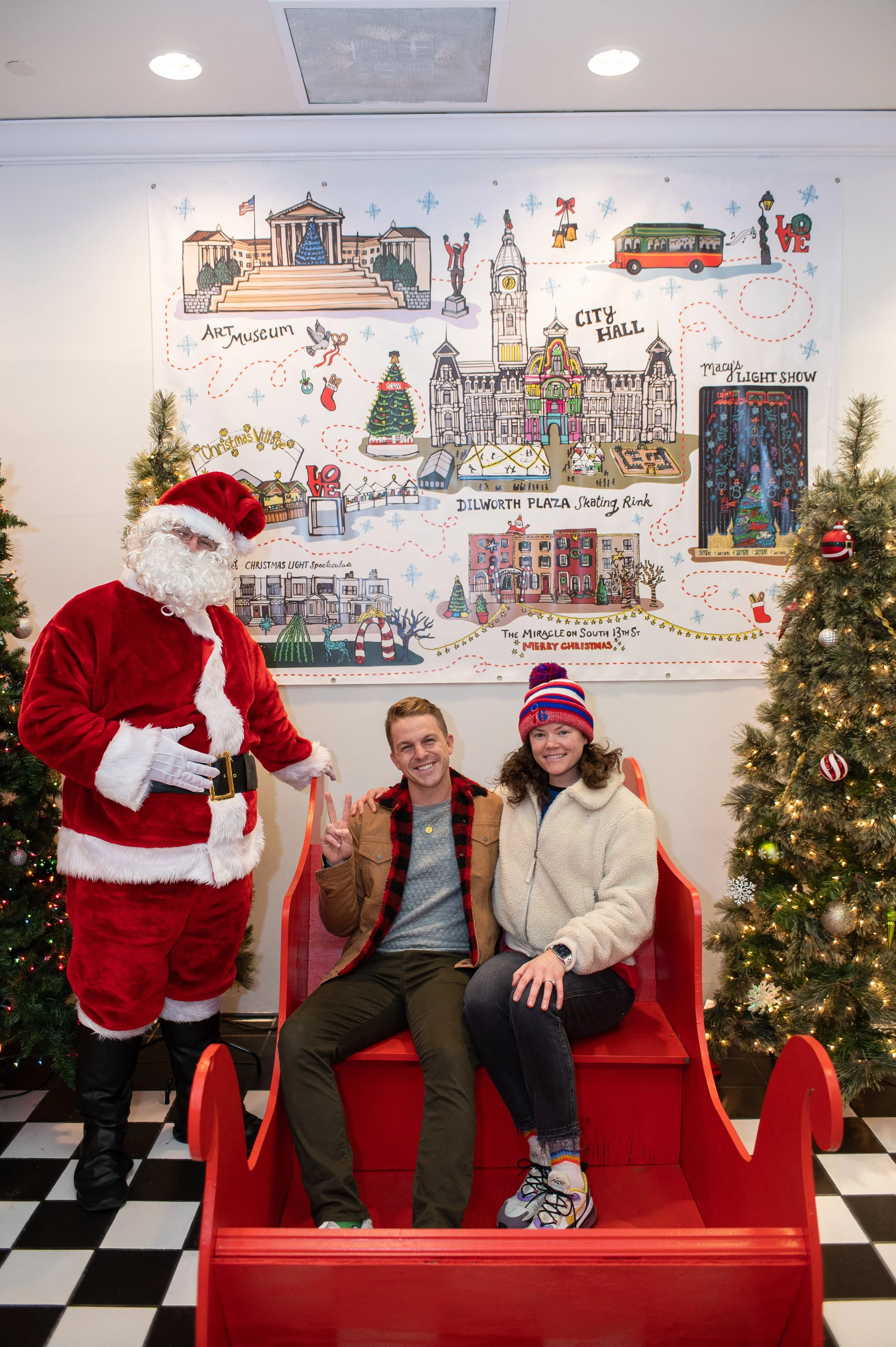 Santa Claus standing next to two smiling people sitting on a red sleigh bench, decorated for Christmas, with two Christmas trees on either side. The background features a Christmas-themed illustrated map of a city.