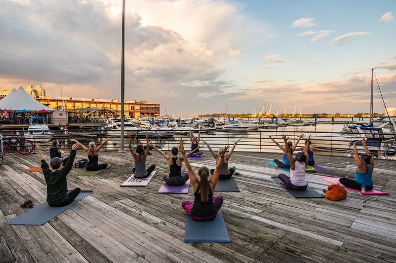 Group of people participating in outdoor yoga class on wooden dock near marina, overlooking boats and water at sunset.