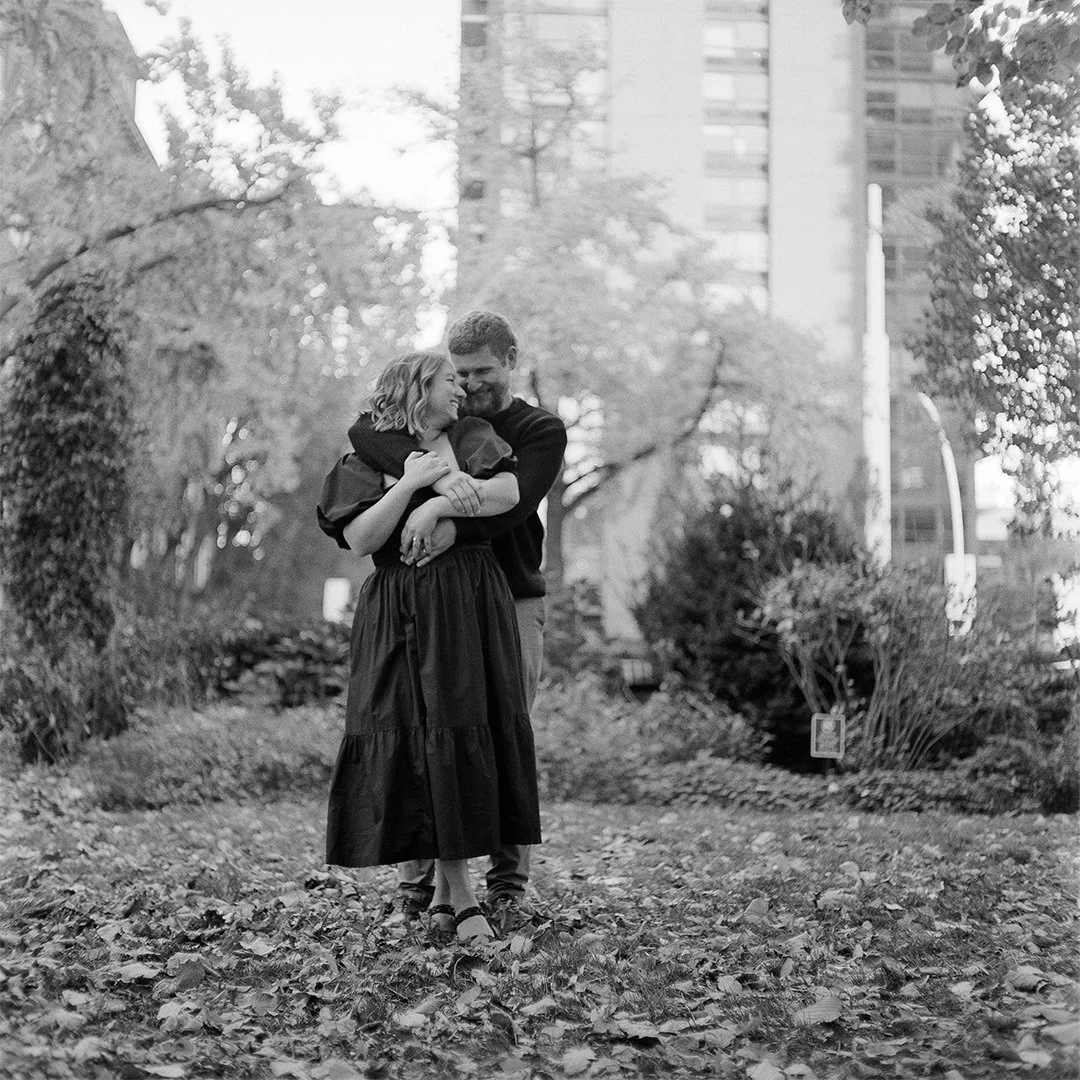 A black and white photo of a couple hugging and smiling in a park with autumn leaves on the ground, trees, and tall buildings in the background.