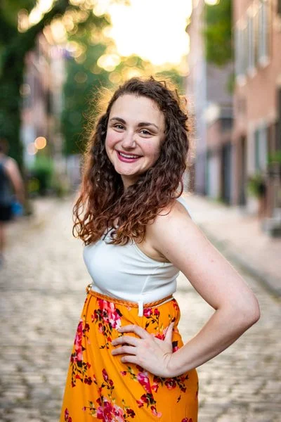 Young woman with curly brown hair smiling outdoors in a street with buildings and trees in the background, wearing a white top and a yellow floral skirt.