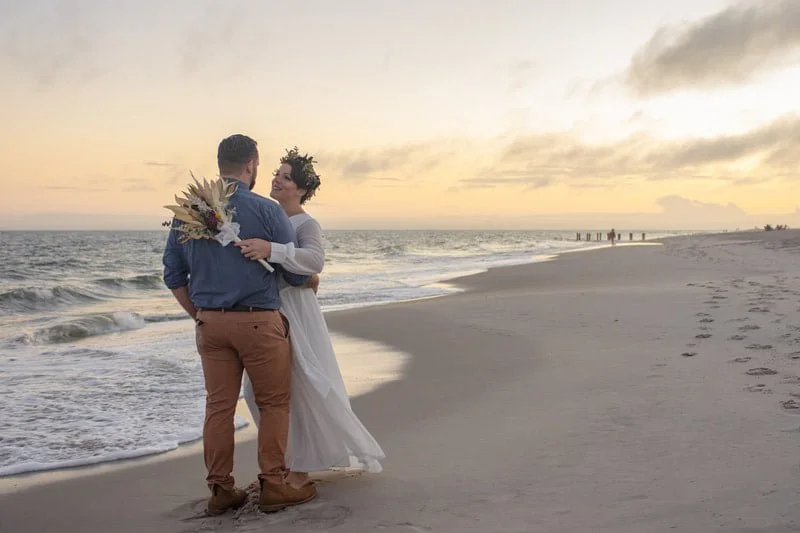 A couple standing on the beach at sunset, embracing, with a bouquet of flowers, the ocean in the background.