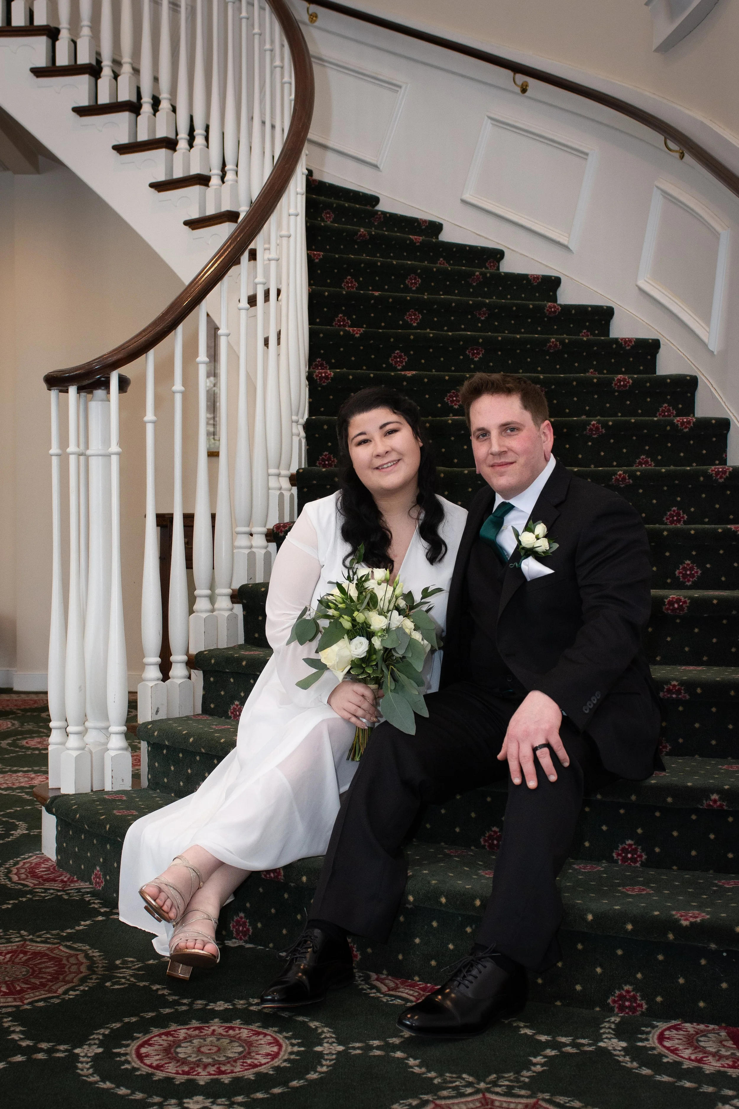 A newlywed couple sitting on a staircase, smiling at the camera. The bride is wearing a white dress, holding a bouquet of white flowers, and the groom is dressed in a black tuxedo with a green tie and a white boutonniere.