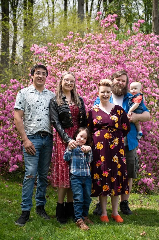 Family standing outdoors in front of pink flowering bushes, with four adults and one child.