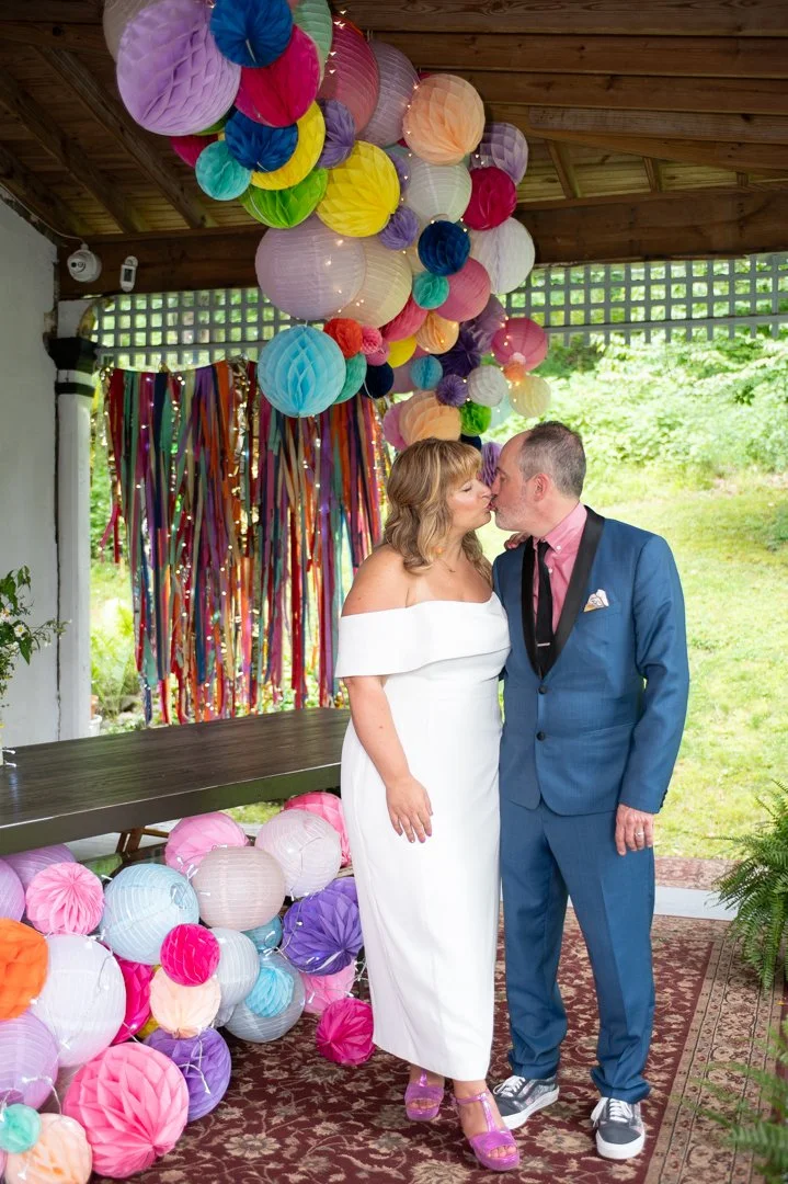 A couple dressed in wedding attire sharing a kiss at a celebration decorated with colorful paper lanterns and streamers.