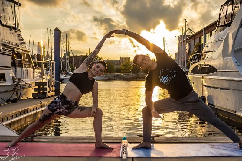 A woman and man practicing yoga on a dock at sunset, forming a heart shape with their arms, with boats and a harbor in the background.