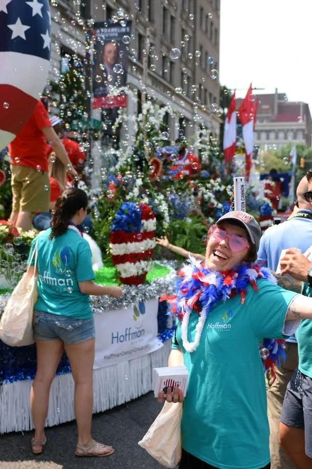 People celebrating in a parade with a float decorated with red, white, and blue flowers, American flags, and Christmas trees, with one woman smiling and wearing sunglasses and a hat, holding a small booklet.