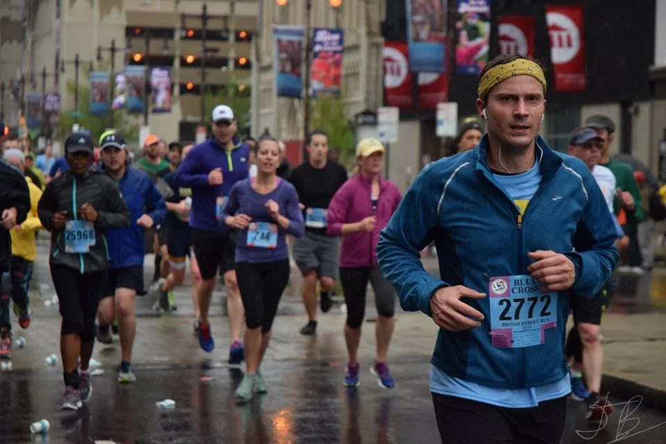 People running in a marathon on a wet city street, with buildings and colorful banners in the background.