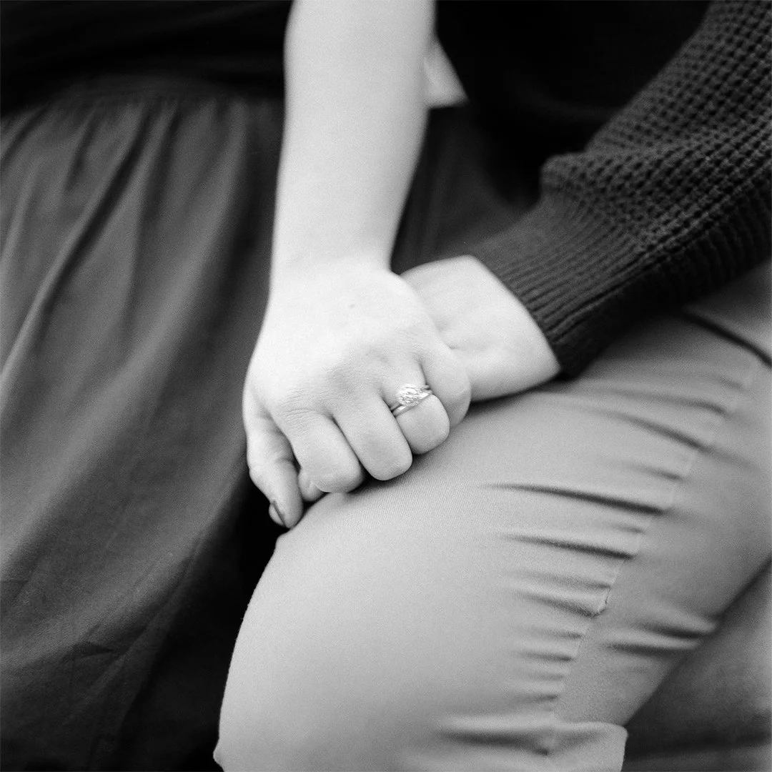 Close-up of two people holding hands; one person's hand with a ring resting on the other person's thigh.