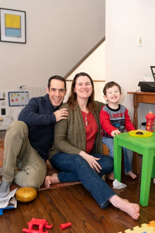Three people, likely a family, smiling and sitting on the floor in a living room with a staircase in the background. A man and woman are sitting close together, and a young boy is sitting on a small green table with toys around.