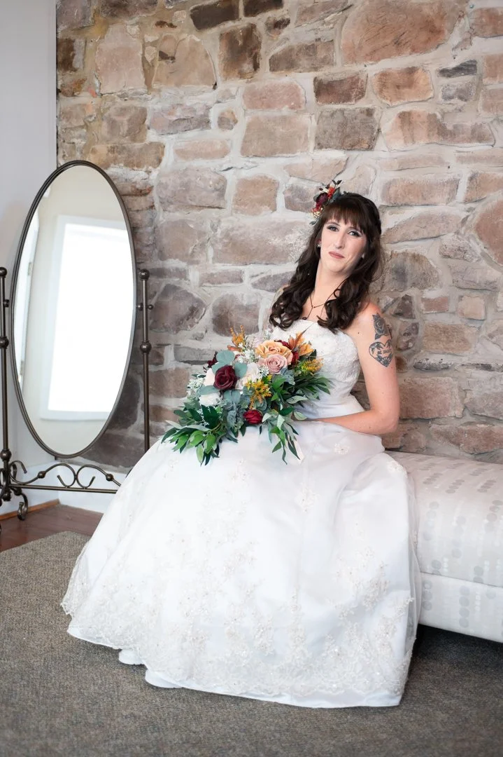 A bride in a white wedding gown sitting on a cushioned bench in front of a rustic brick wall, holding a colorful bouquet of flowers, with a large mirror nearby reflecting a window.