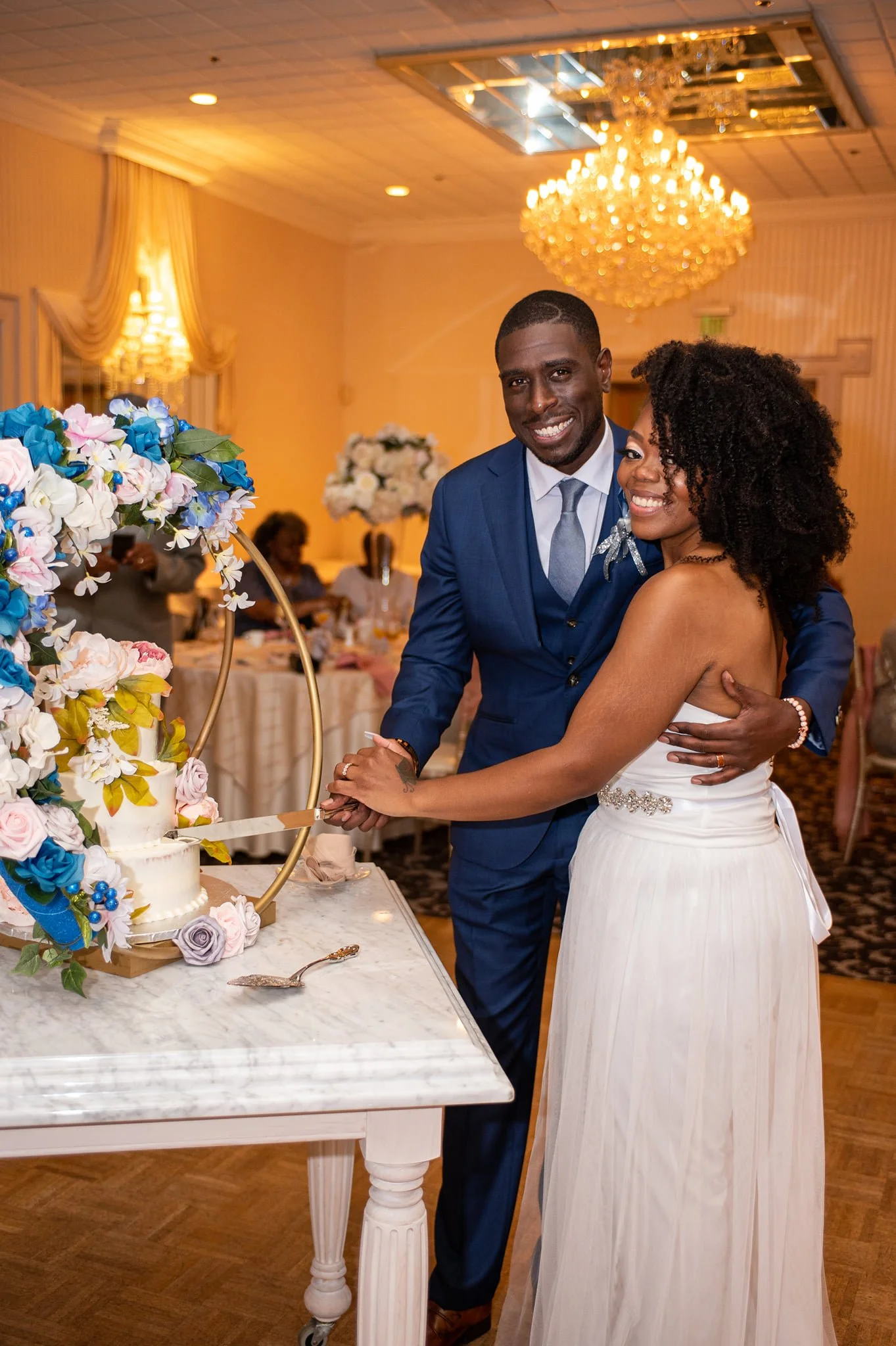 A newlywed couple cutting their wedding cake together at their reception. The groom is wearing a blue suit and the bride is in a white strapless dress. There are floral decorations and a chandelier overhead.
