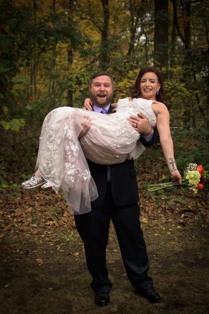 A man in a black suit and purple tie is smiling and holding a woman in a wedding dress, who is carrying a bouquet, in a wooded outdoor setting.