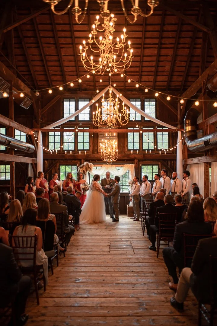 Wedding ceremony inside a rustic wooden barn with chandeliers and string lights, with the bride and groom at the altar surrounded by bridal party and guests.