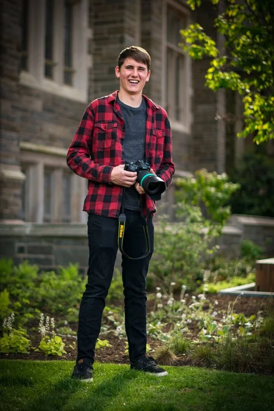 A young man in a red and black plaid shirt holding a DSLR camera, standing on a well-maintained garden lawn in front of a historic stone building with greenery.