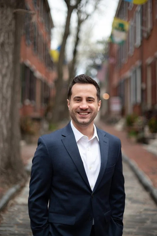 A smiling man in a navy blue suit and white shirt standing on a city sidewalk with trees and residential buildings in the background.
