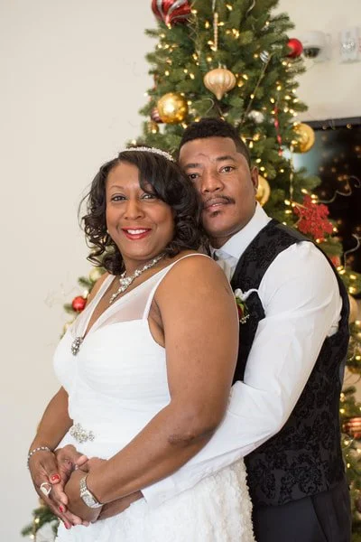 A woman in a white dress and a man in a suit with a vest, standing in front of a decorated Christmas tree.