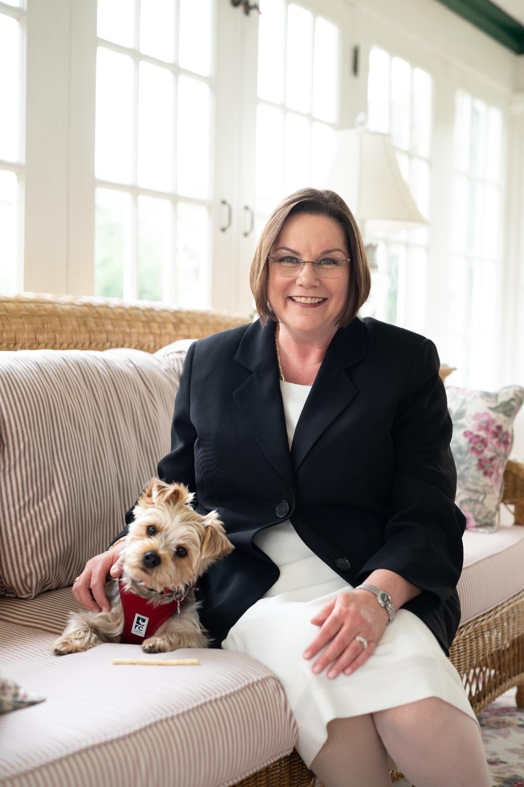 A woman with glasses and a bob haircut, wearing a black blazer and white dress, sitting on a striped sofa with a small dog wearing a red harness, in a sunlit room with large windows and white walls.