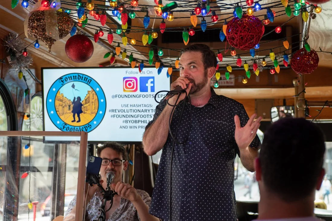 A man with a beard and short dark hair singing into a microphone during a festive gathering. Behind him, there is another man with glasses playing an instrument. The setting is decorated with colorful string lights and red and gold ornaments. A screen displaying social media information and a logo for 'Founding Footsteps' is visible in the background.