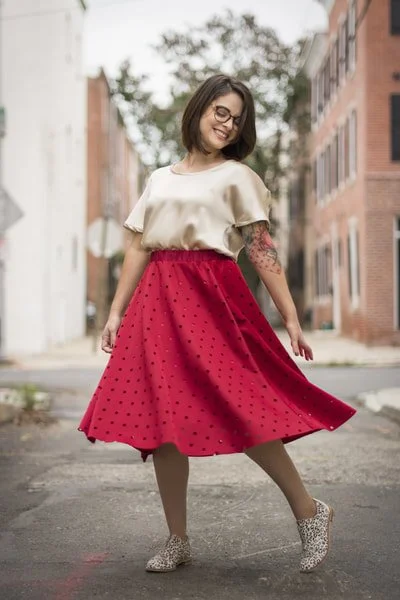 A woman with glasses and a floral tattoo on her arm, wearing a beige top and a red, polka-dot skirt, stands on a street with brick buildings.