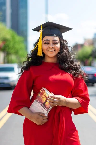 Young woman in a red graduation gown and cap holding a diploma outdoors with city buildings and cars in the background.