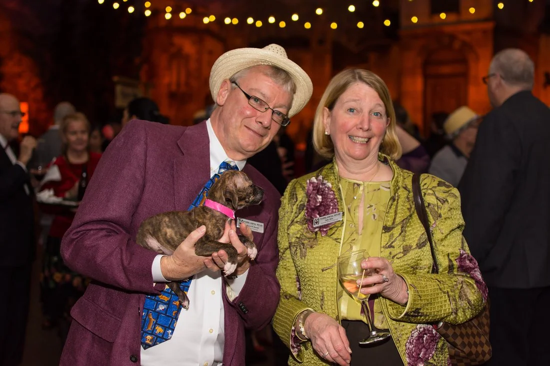 A man and a woman are smiling at a social event. The man is holding a small puppy and has a name tag, wearing a hat, glasses, and a suit. The woman is holding a glass of wine, has a name tag, and is dressed in a yellow patterned jacket. There are other people in the background, with warm lighting and wooden decor.