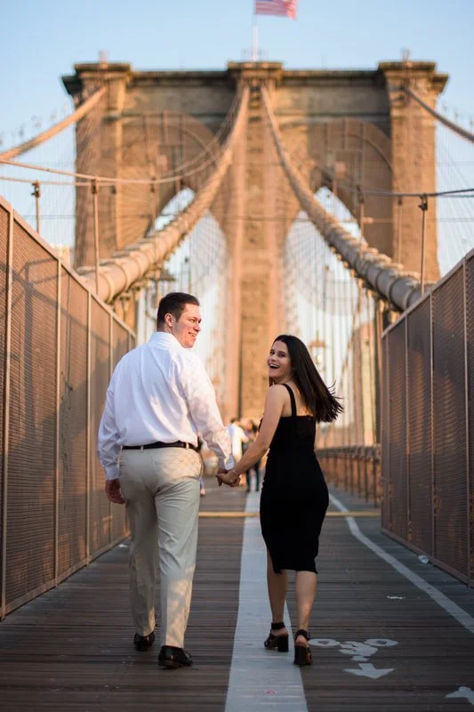 Couple holding hands and walking on Brooklyn Bridge during sunset.